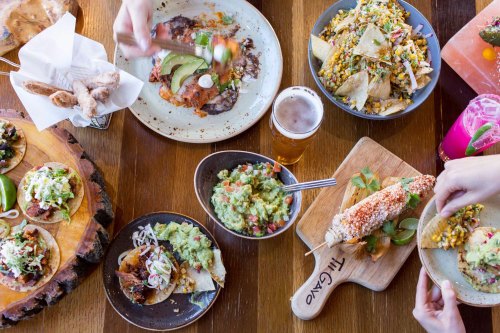 A vibrant spread of tacos, nachos, guacamole, corn, and drinks on a wooden table, showcasing a festive meal.