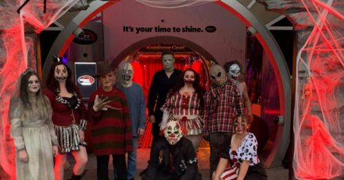 A group of people in Halloween costumes poses in a spooky, decorated setting with red lighting and cobwebs.