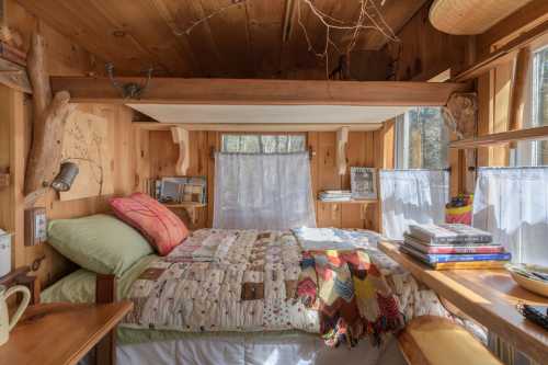 Cozy wooden bedroom with a bed, colorful quilt, books, and large windows letting in natural light.