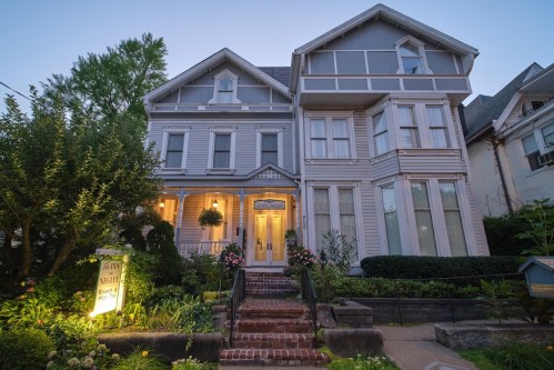 A charming Victorian-style house with a blue exterior, lush gardens, and a welcoming entrance at dusk.