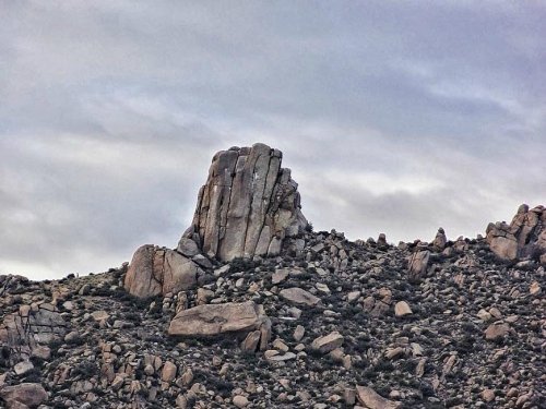 A rugged rock formation rises above a rocky landscape under a cloudy sky.