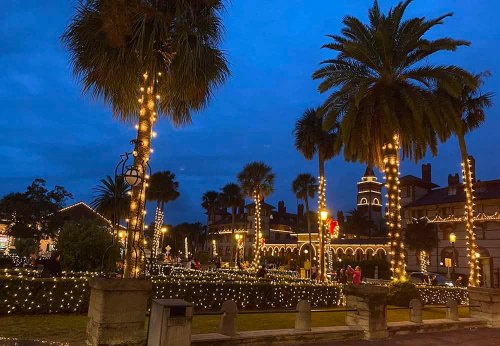 A festive scene with palm trees and buildings adorned with twinkling lights under a twilight sky.