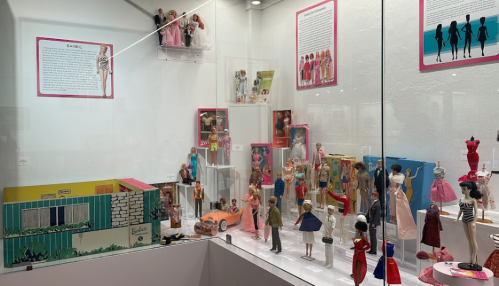 Display case featuring a collection of vintage Barbie dolls, outfits, and accessories in a museum setting.