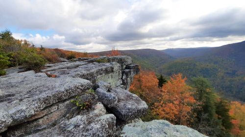 A rocky cliff overlooks a colorful autumn landscape with trees and rolling hills under a cloudy sky.