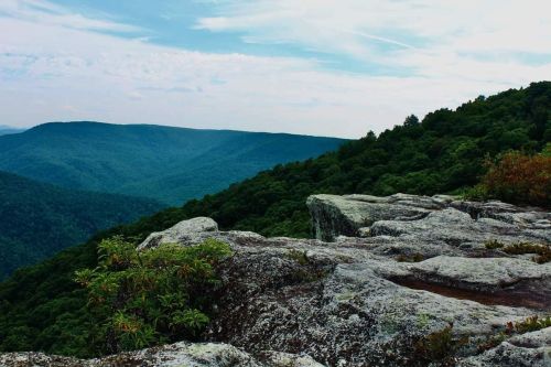 A rocky outcrop overlooks a lush green valley and distant mountains under a cloudy sky.