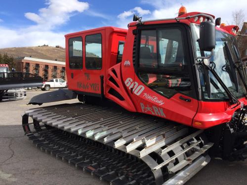 A red snowcat vehicle with a cabin and metal tracks, parked in a snowy area with buildings in the background.