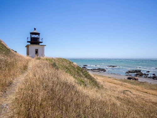 A lighthouse stands on a grassy hill overlooking a rocky beach and the ocean under a clear blue sky.
