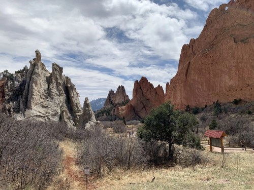 Rocky formations rise against a cloudy sky, with sparse vegetation and a wooden sign in a natural landscape.