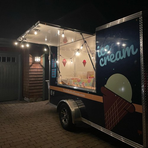 A brightly lit ice cream truck parked in a driveway, featuring colorful ice cream cones on the side.