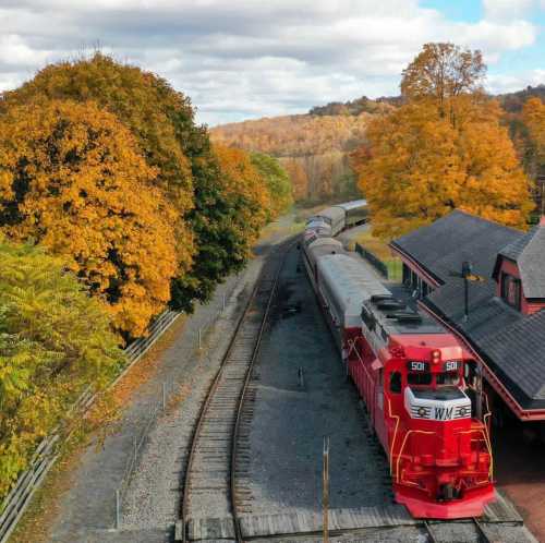 A train on a curved track near a station, surrounded by vibrant autumn foliage and rolling hills.