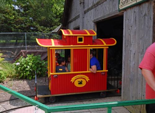 A colorful red train ride with passengers, parked outside a wooden building in a park setting.