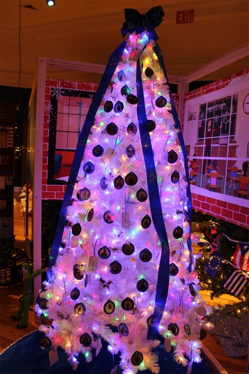 A brightly lit white Christmas tree adorned with black ornaments and blue ribbons, set against a festive backdrop.