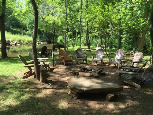 A serene outdoor setting with wooden chairs arranged around a fire pit near a creek, surrounded by lush greenery.