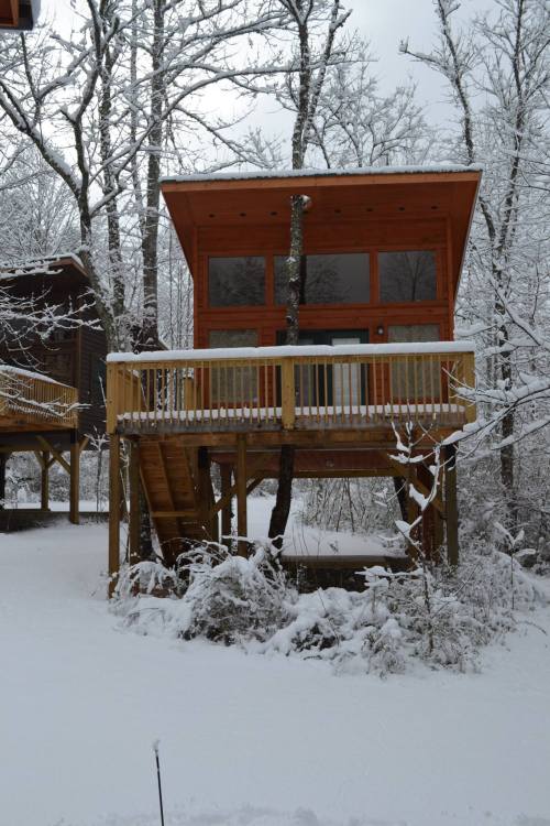 A modern cabin on stilts surrounded by snow-covered trees, featuring large windows and a wooden deck.