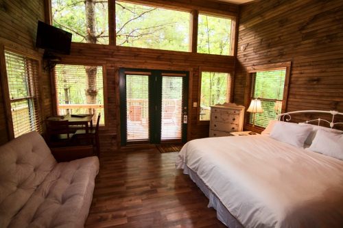 Cozy wooden cabin interior with a bed, sofa, and large windows overlooking greenery.