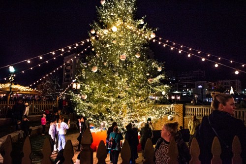 A festive Christmas tree adorned with lights and ornaments, surrounded by people enjoying the holiday atmosphere at night.