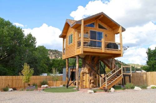 A wooden treehouse with a balcony, elevated on stilts, surrounded by trees and a fenced yard.