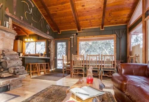 Cozy cabin interior with wooden beams, a stone fireplace, and rustic furniture, featuring a dining area and large windows.
