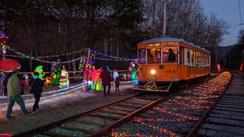 A brightly lit orange train surrounded by festive decorations and people enjoying a holiday display at night.