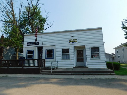 A small white building with a porch, featuring a U.S. Post Office sign and an American flag outside.