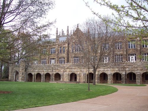 Historic stone building with arched windows, surrounded by trees and a grassy area, under a cloudy sky.