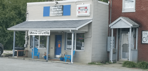 A small storefront with a sign for "Upton Eats And More" and a "Grand Opening" banner, featuring blue chairs outside.