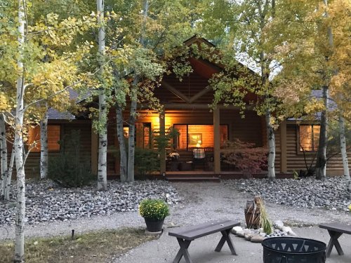 A cozy wooden cabin surrounded by trees, with warm light glowing from the windows and a fire pit in the foreground.