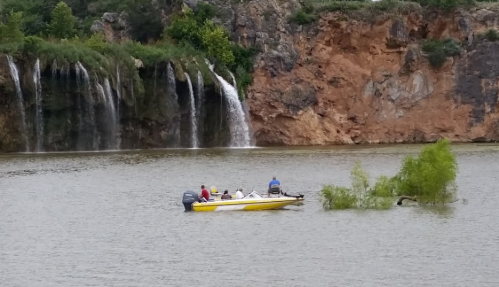 A small boat with four people floats on a lake near a rocky shore and a waterfall in the background.