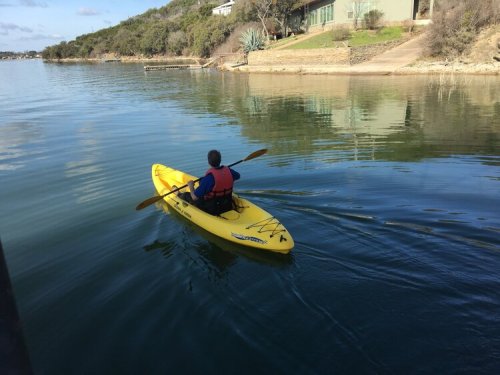 A person in a yellow kayak paddles on calm water near a shoreline with trees and a house in the background.