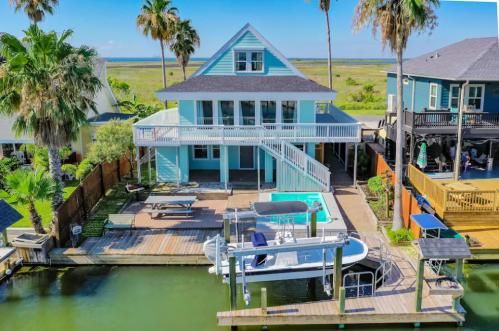 Aerial view of a colorful waterfront house with a pool, surrounded by palm trees and a dock for boats.