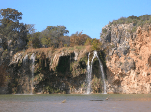 A scenic view of a waterfall cascading down a rocky cliff into a calm body of water, surrounded by trees and blue sky.