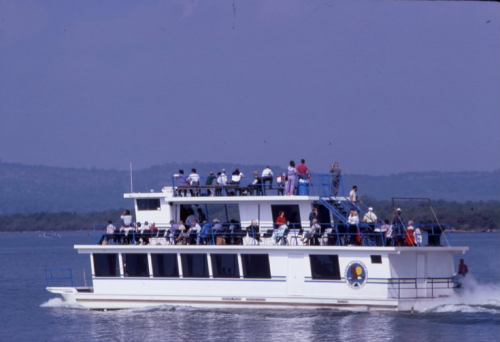 A large white ferry with passengers on multiple decks cruising on a calm body of water against a scenic backdrop.