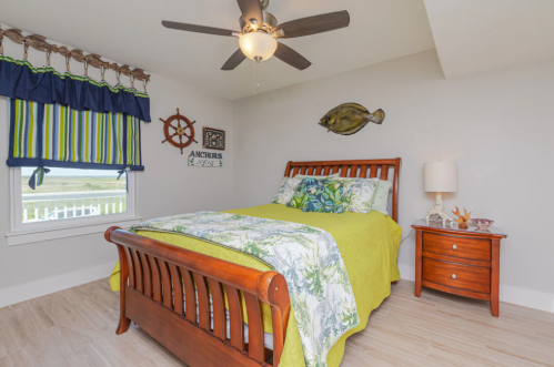 Cozy bedroom with a wooden bed, colorful bedding, nautical decor, and a ceiling fan. Bright and inviting atmosphere.