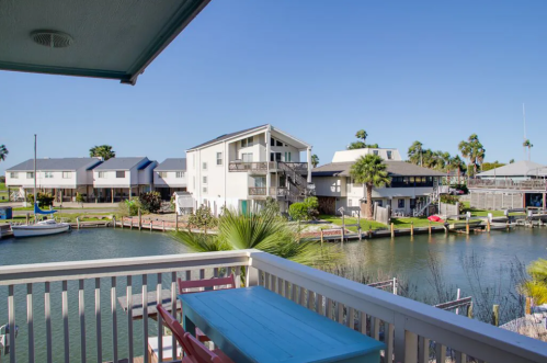 View from a balcony overlooking a canal with boats and houses, surrounded by palm trees under a clear blue sky.