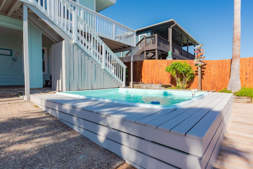 A small, inviting hot tub on a wooden deck, surrounded by a fenced yard and nearby houses under a clear blue sky.