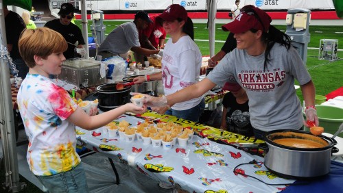A child in a tie-dye shirt receives a food sample from a woman in a gray shirt at a festive outdoor event.