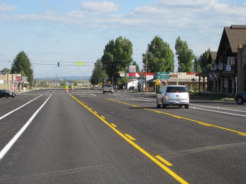 A wide, empty road with a few cars, lined by trees and buildings under a clear blue sky. Traffic lights visible ahead.