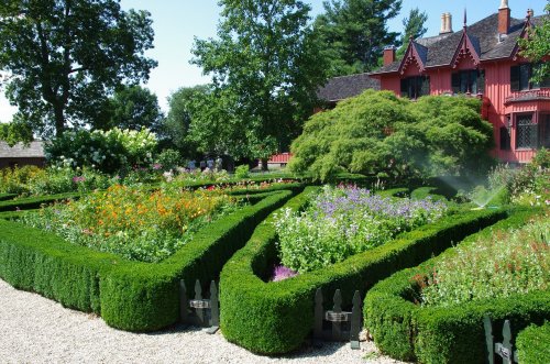 A vibrant garden with neatly trimmed hedges and colorful flowers, set against a red house and blue sky.