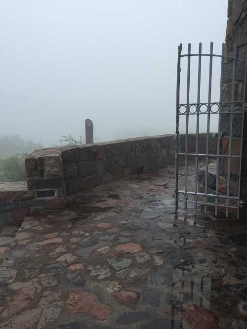 A stone pathway leads to a metal gate, surrounded by thick fog and wet surfaces, creating a mysterious atmosphere.