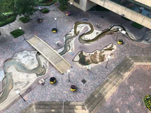 Aerial view of a landscaped area featuring a winding water feature and a bridge, surrounded by planters and brick pathways.