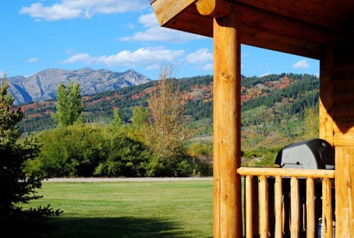 A wooden porch with a grill overlooks a lush green lawn and mountains in the background under a blue sky.