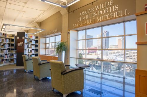 Interior of a library featuring bookshelves, seating area, and large windows with city views. Names of authors on the wall.