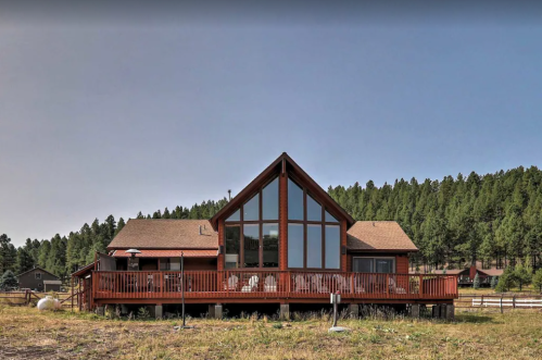 A large wooden house with a triangular roof and large windows, surrounded by trees and open land.