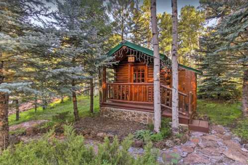 A cozy wooden cabin surrounded by tall trees and greenery, featuring a small porch and stone pathway.