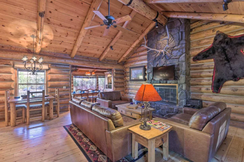 Cozy log cabin interior featuring a stone fireplace, leather sofas, wooden dining table, and rustic decor.