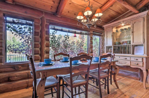 Cozy log cabin dining area with a wooden table, chairs, and a chandelier, featuring large windows with scenic views.