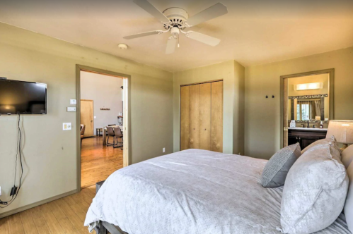 A cozy bedroom with a bed, ceiling fan, TV, and a doorway leading to another room. Light-colored walls and wooden floors.
