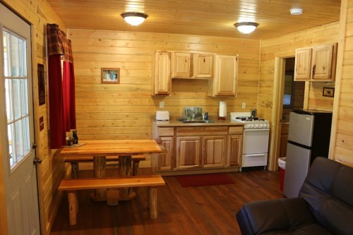 Cozy wooden cabin interior featuring a kitchen, dining table, and living area with a sofa. Warm, rustic decor.