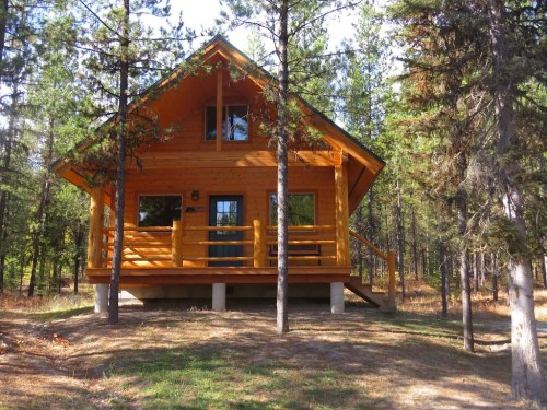 A wooden cabin surrounded by tall trees, featuring a porch and large windows, set in a natural forested area.