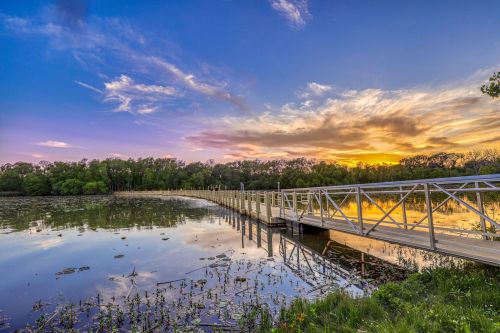 A wooden pier extends into a calm lake at sunset, surrounded by lush greenery and colorful clouds.